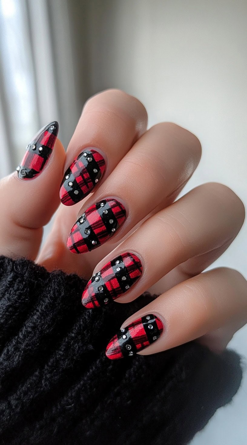 Almond-shaped nails with a bold red-and-black plaid pattern accented by black rhinestone-studded bands.