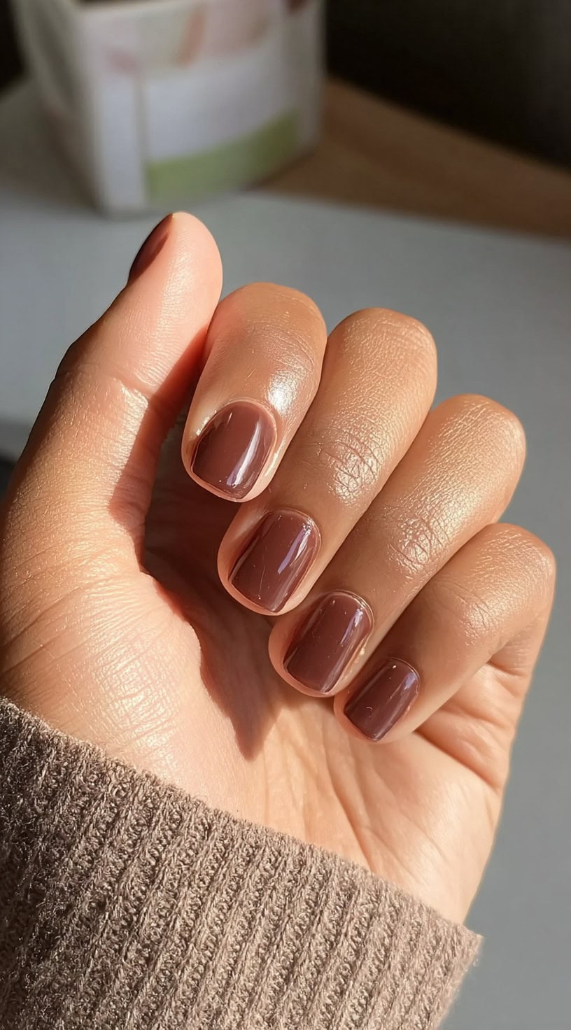 Short, rounded chocolate brown nails on a hand with medium brown skin tone, glowing in sunlight.