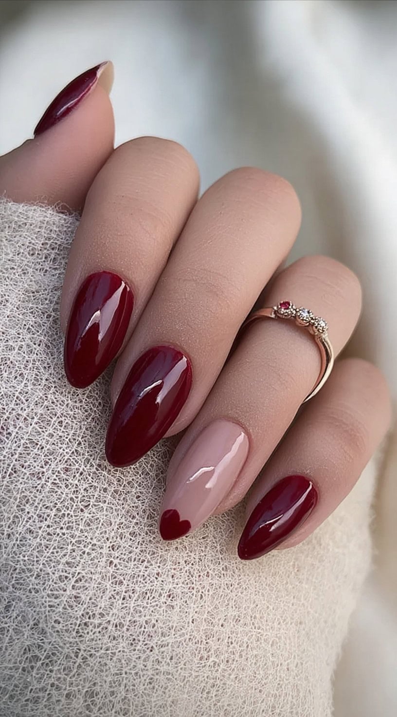 Almond-shaped nails painted in a glossy wine red with one nude accent nail featuring a tiny red heart at the tip.