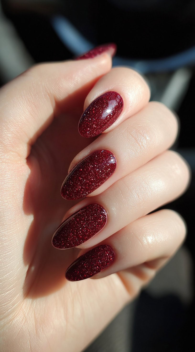 A hand with uniformly shaped almond nails fully covered in wine red glitter under a high-shine topcoat.