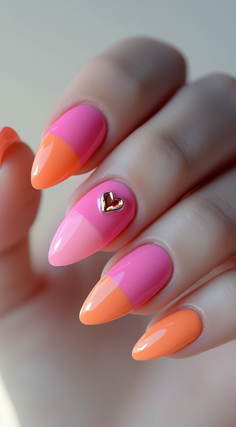 Almond-shaped nails with a half-and-half split of orange and pink polish, accented with a metallic heart charm.