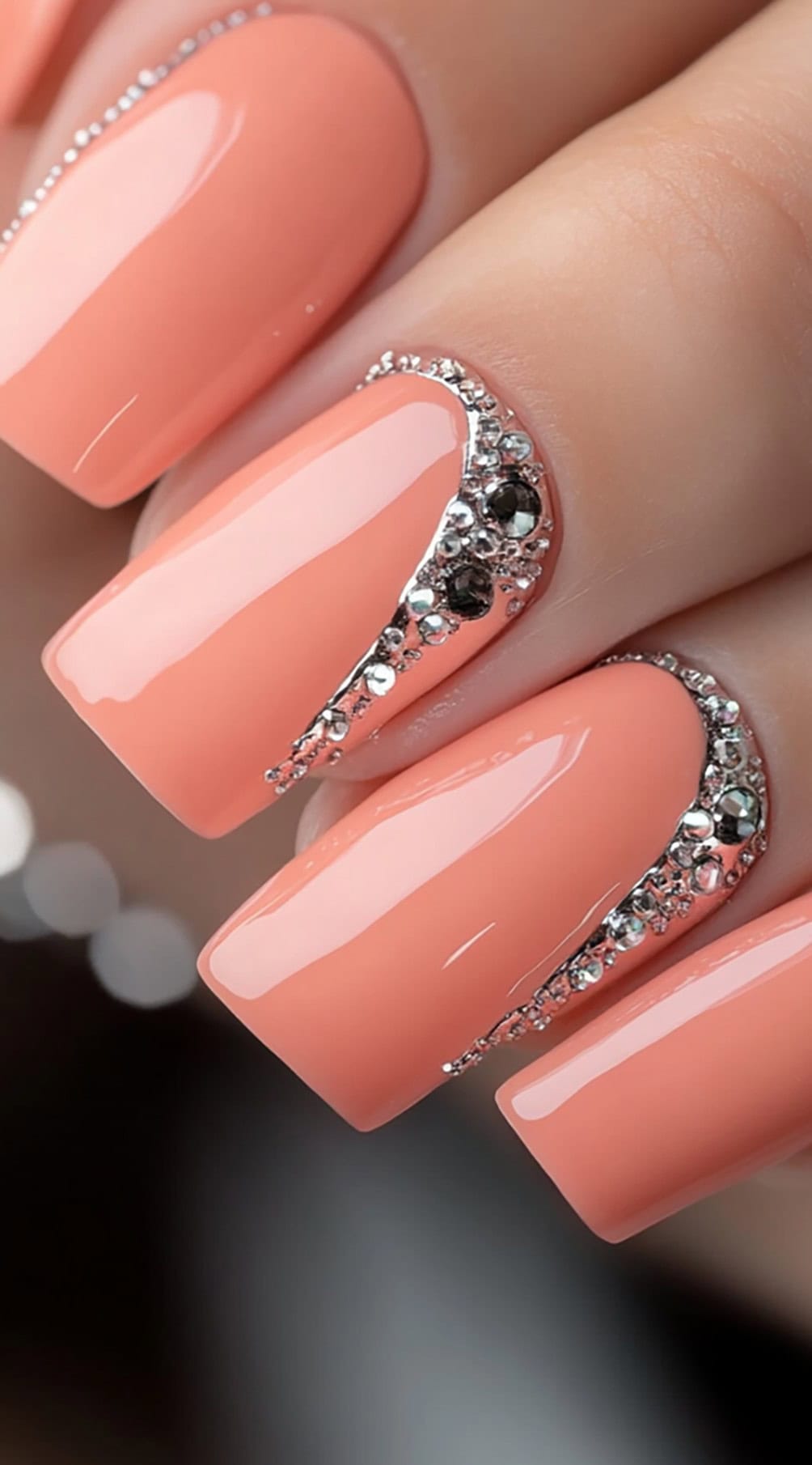 A set of square-shaped nails in a warm coral pink shade, adorned with silver and black rhinestone embellishments near the cuticle.