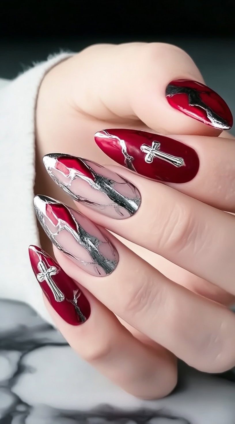 Almond-shaped nails combining deep red polish, silver metallic veining, and raised silver cross embellishments.