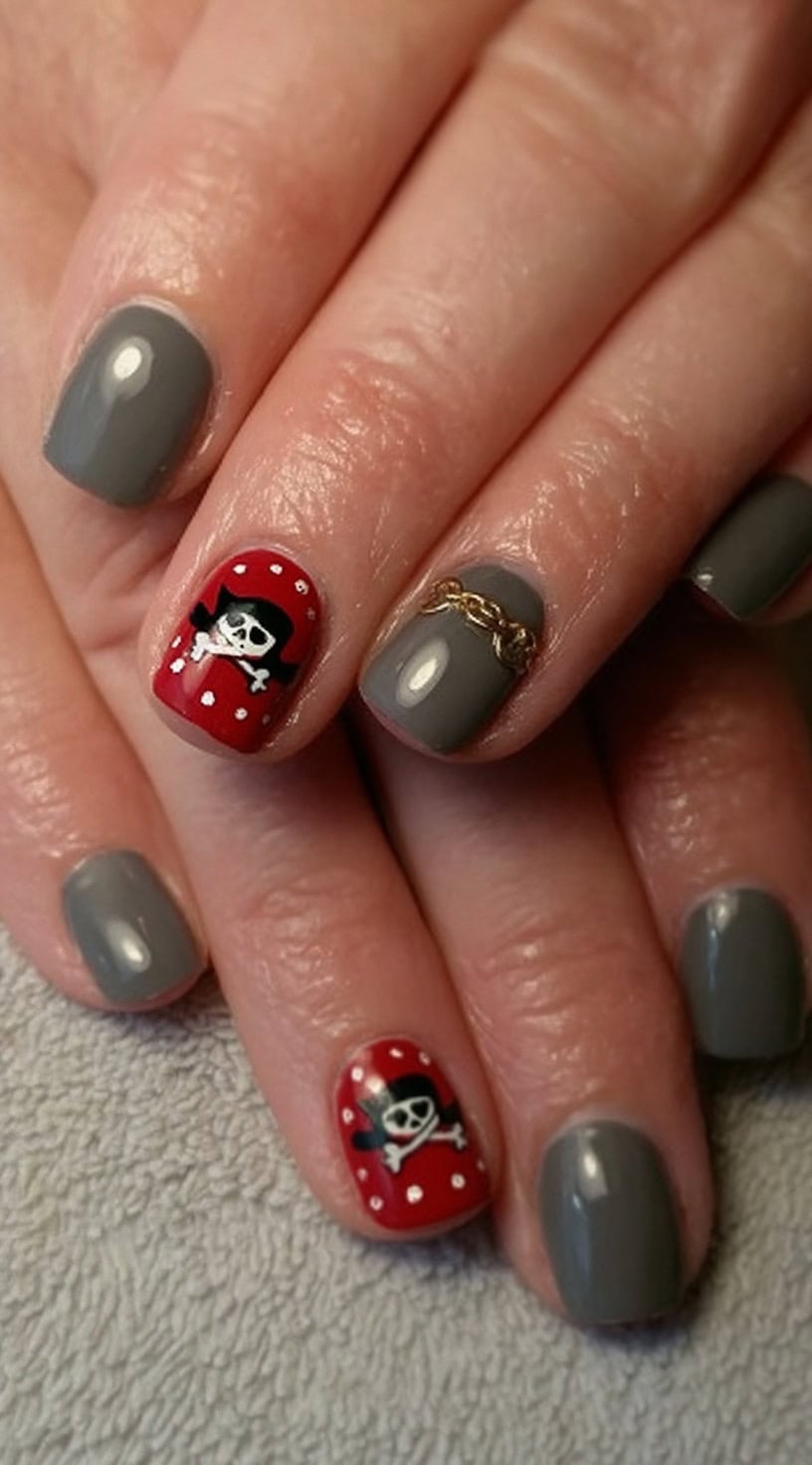 Short grey nails are accented with red polka dot pirate skulls and a small gold chain on one nail.