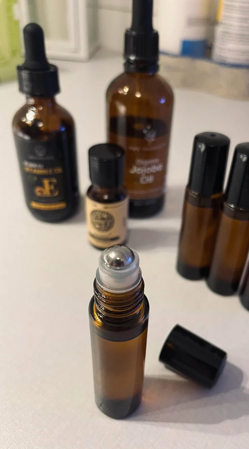 A close-up of a filled amber glass roller bottle for cuticle oil, surrounded by carrier oils and essential oils.