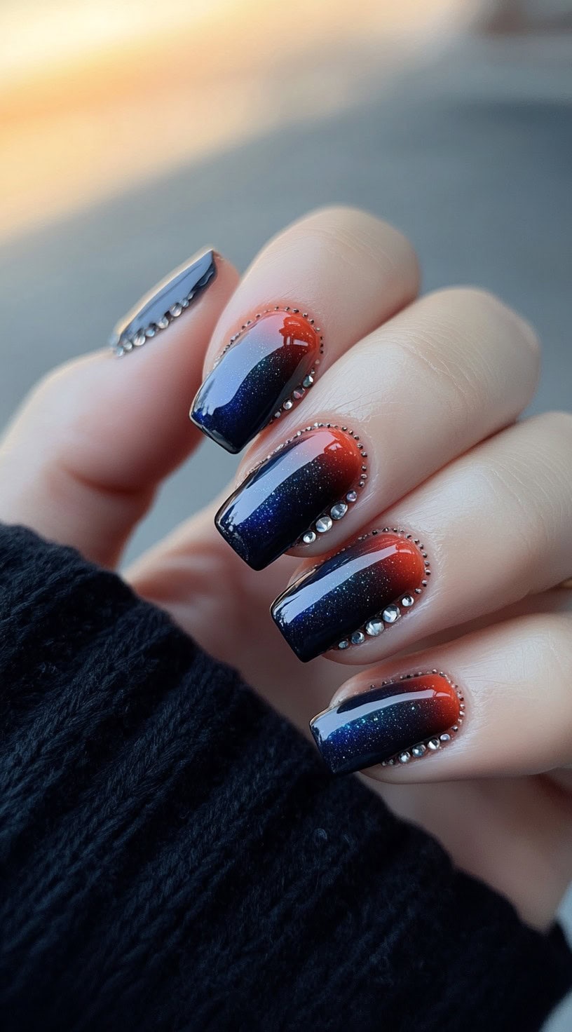 A hand with square-shaped nails featuring a dark ombre gradient transitioning from deep red to navy blue with tiny rhinestones along the edges.