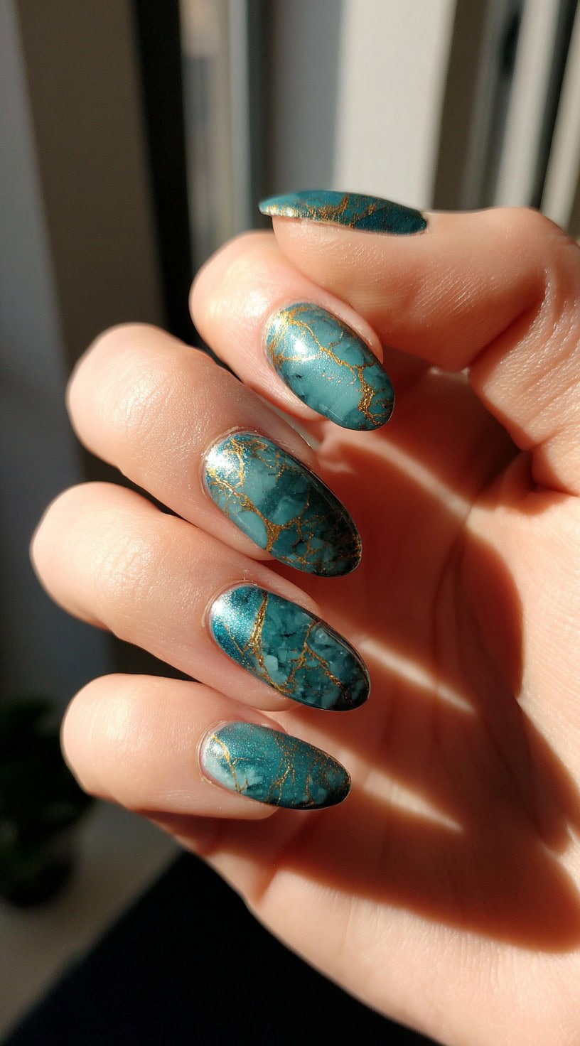 A hand with short oval nails painted in a deep jade teal tone with fine golden marble veining throughout.