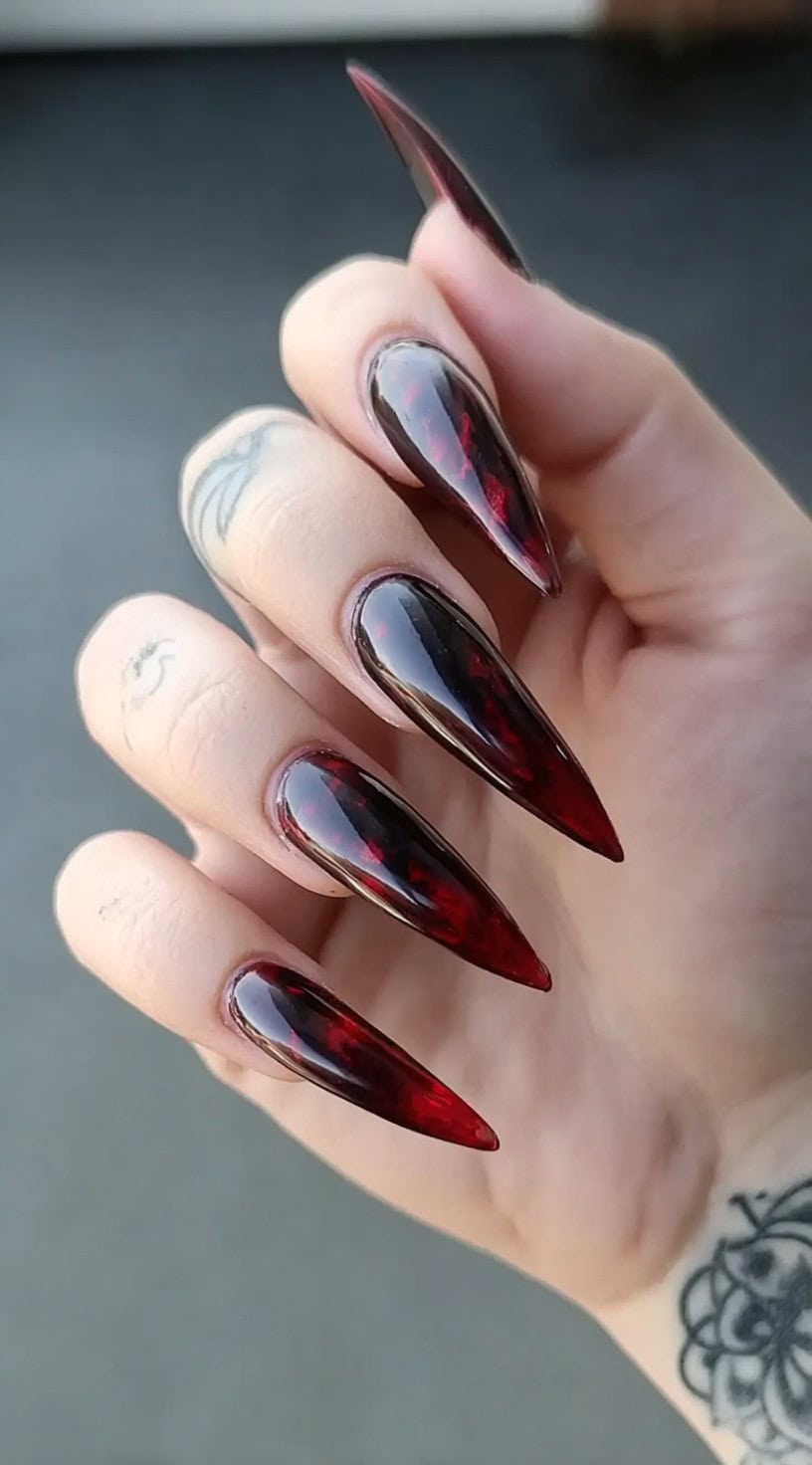 Long, sharply pointed stiletto nails with a glassy finish blending deep red and black hues.
