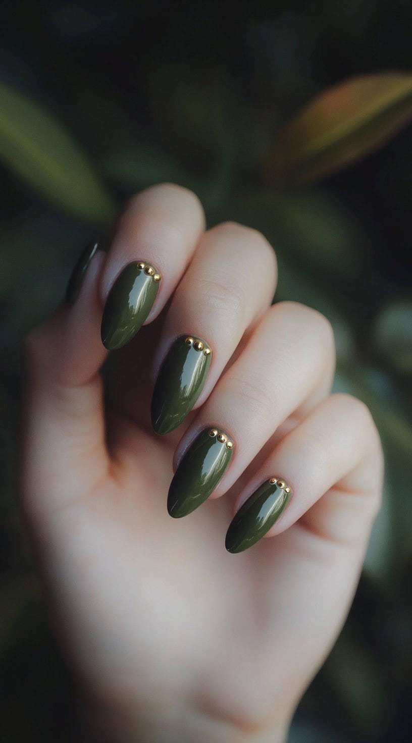 A set of almond-shaped nails painted in a rich deep green shade, adorned with small gold studs at the base for a sleek and modern look.