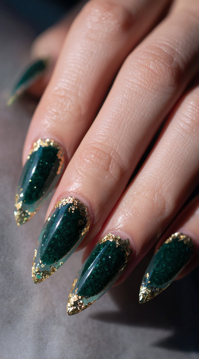 A hand with almond-shaped nails featuring deep green textured polish and jagged gold foil detailing on the tips.