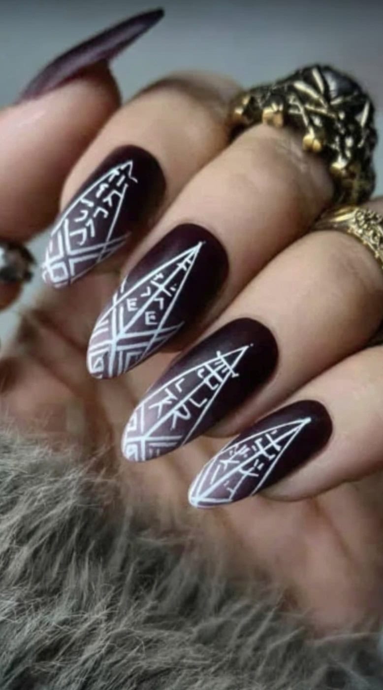A hand with pointed maroon nails featuring sharp white geometric rune designs that form angular symbols.