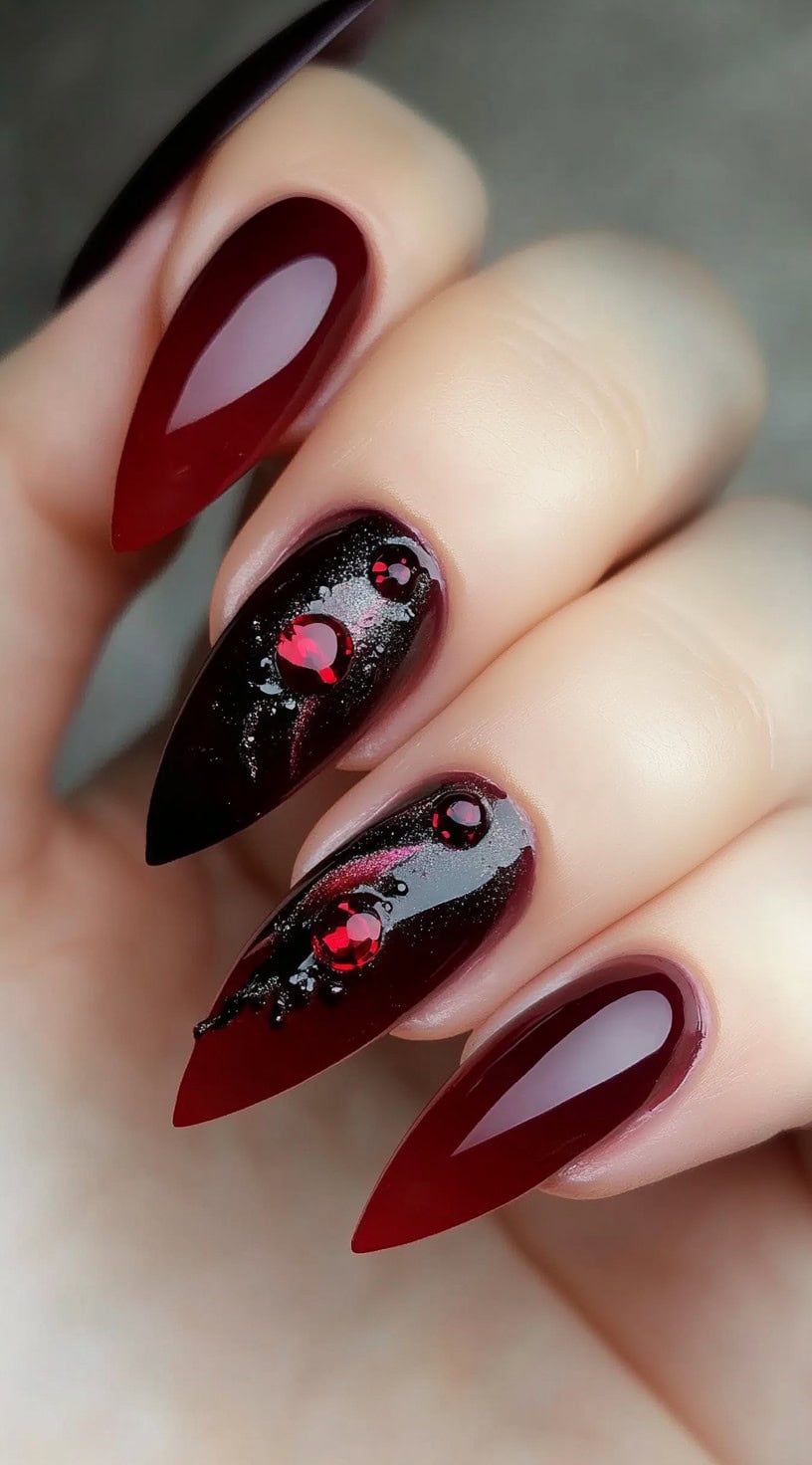 Stiletto-shaped deep oxblood nails with glossy finish, featuring black shimmer and red gem accents on two fingers.