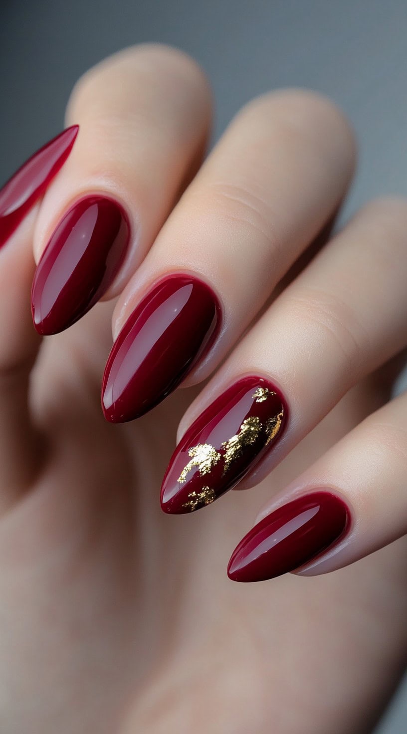 Close-up of almond-shaped deep red nails with a glossy finish and gold foil detail on one nail.