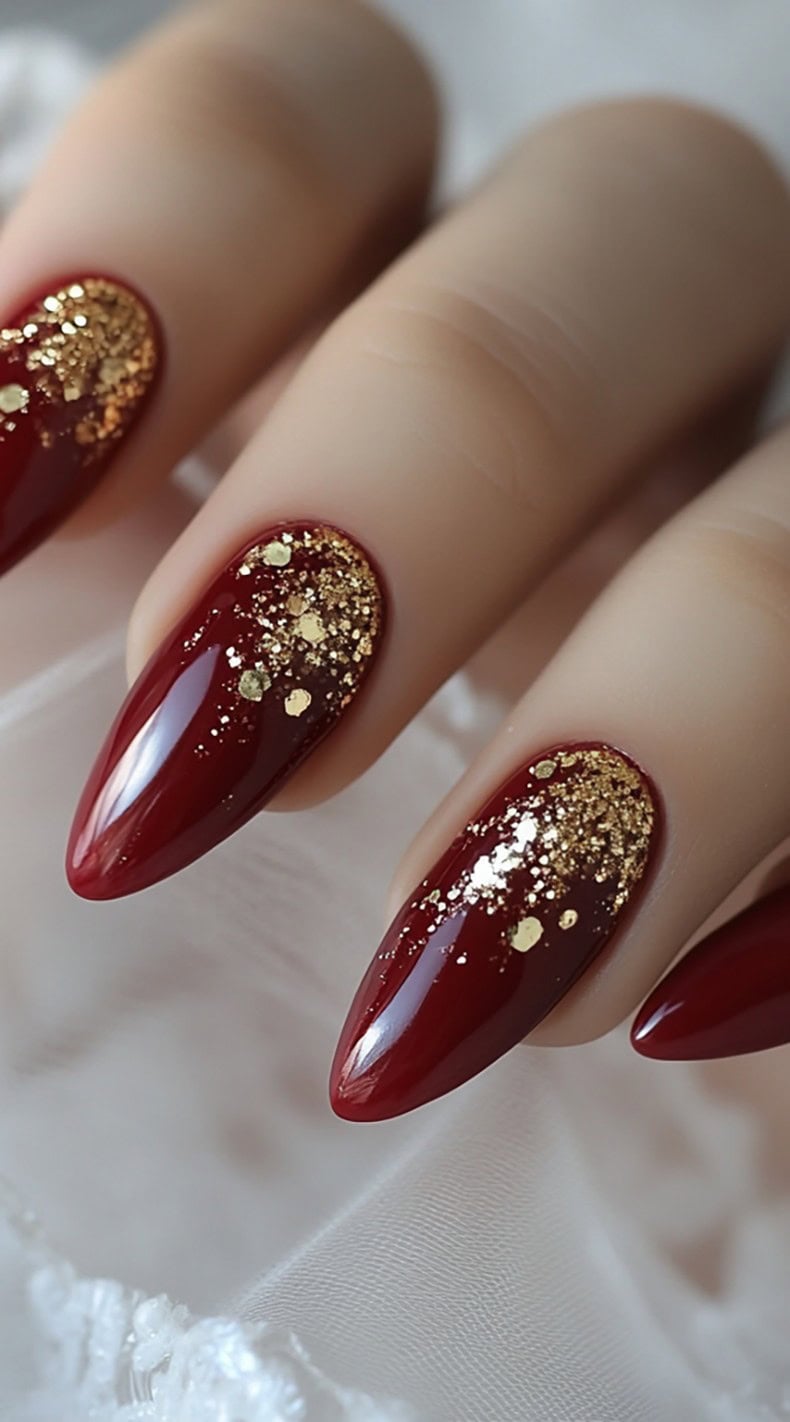 A set of almond-shaped deep red nails featuring gold glitter accents concentrated at the tips, creating a soft, gradient sparkle effect.