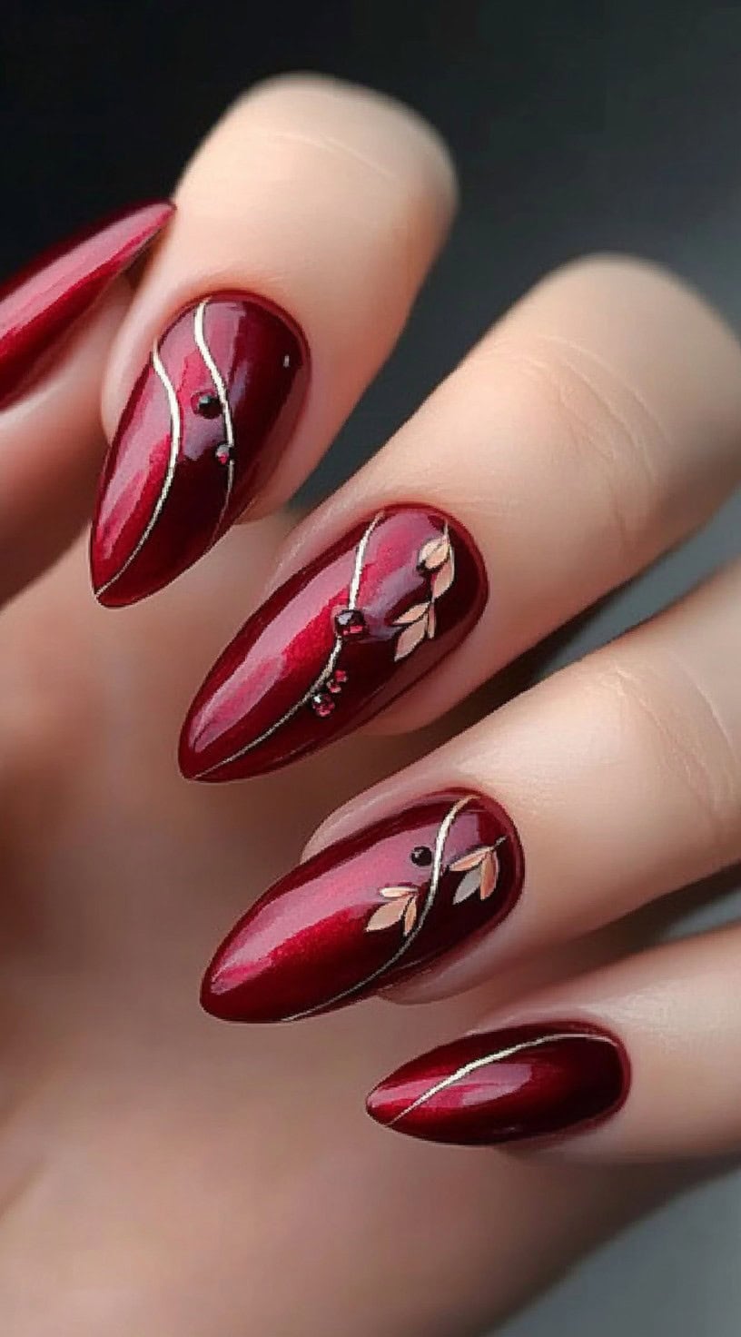 Glossy deep red almond nails featuring delicate silver vines and soft leaf details with small jewel accents.