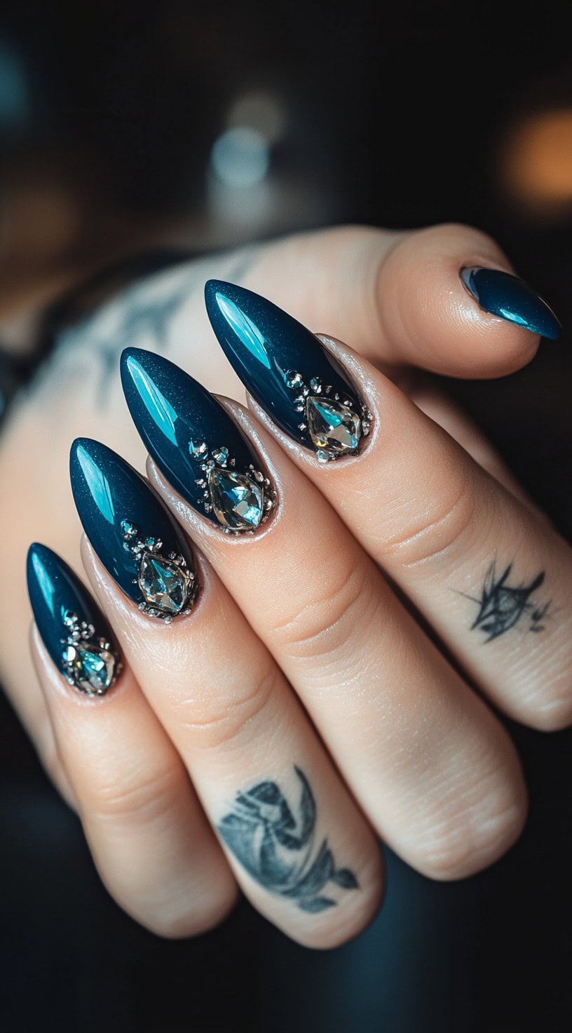 Deep teal stiletto nails adorned with large teardrop-shaped crystals and smaller metallic embellishments at the cuticles.