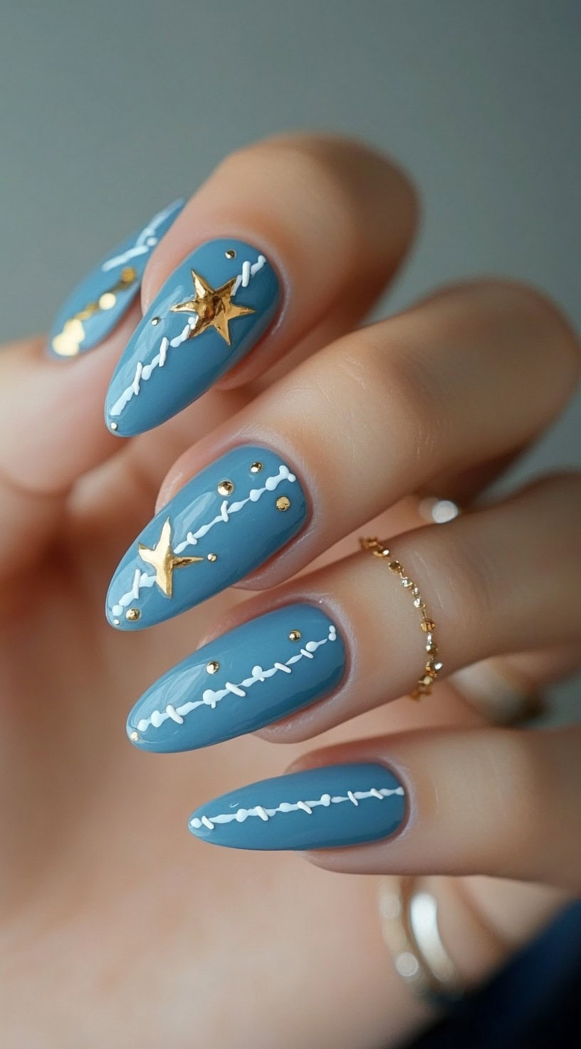 Almond-shaped nails painted in denim blue, featuring gold star embellishments and white stitching details.