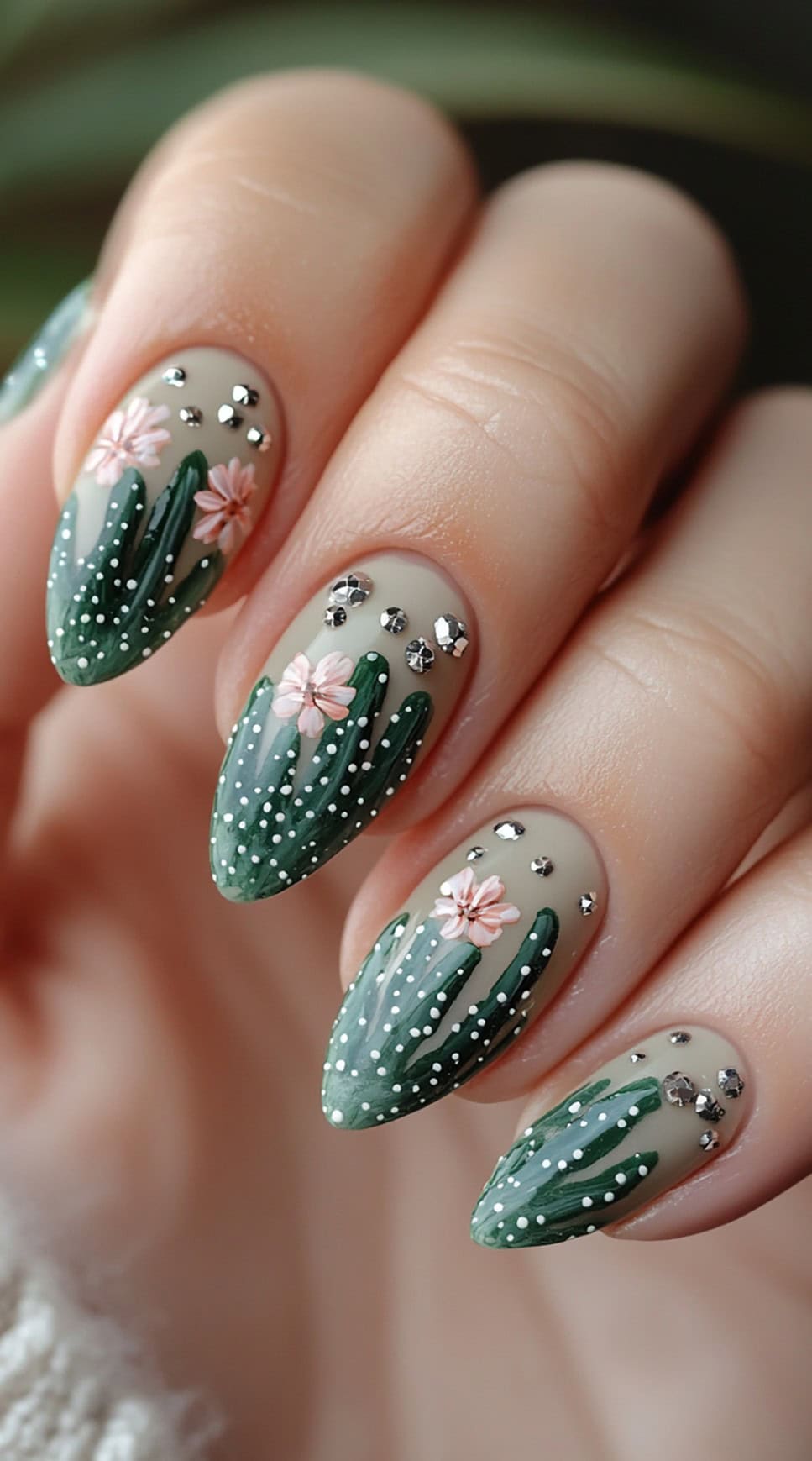 Almond-shaped nails with a muted desert beige base, featuring hand-painted green cacti, tiny pink flowers, and rhinestone accents.