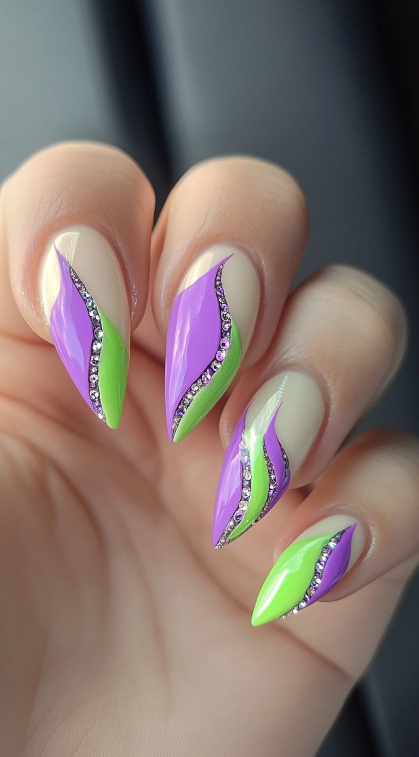 Almond-shaped nails with a creamy base, overlaid with curved ribbons of neon green and lavender purple separated by sparkling rhinestones.