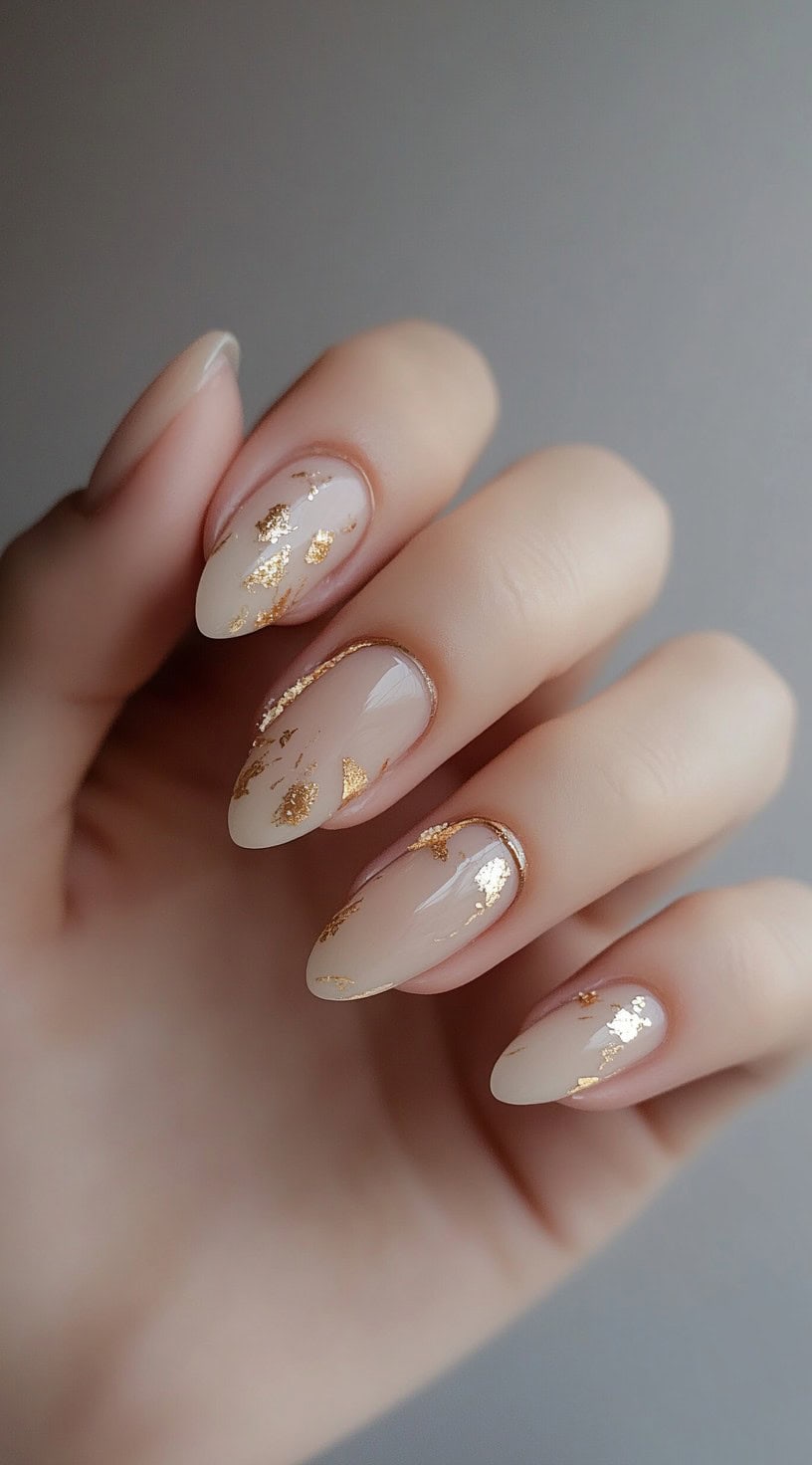 A close-up of a hand featuring soft almond-shaped nails with delicate gold foil accents, creating a refined and luxurious look.