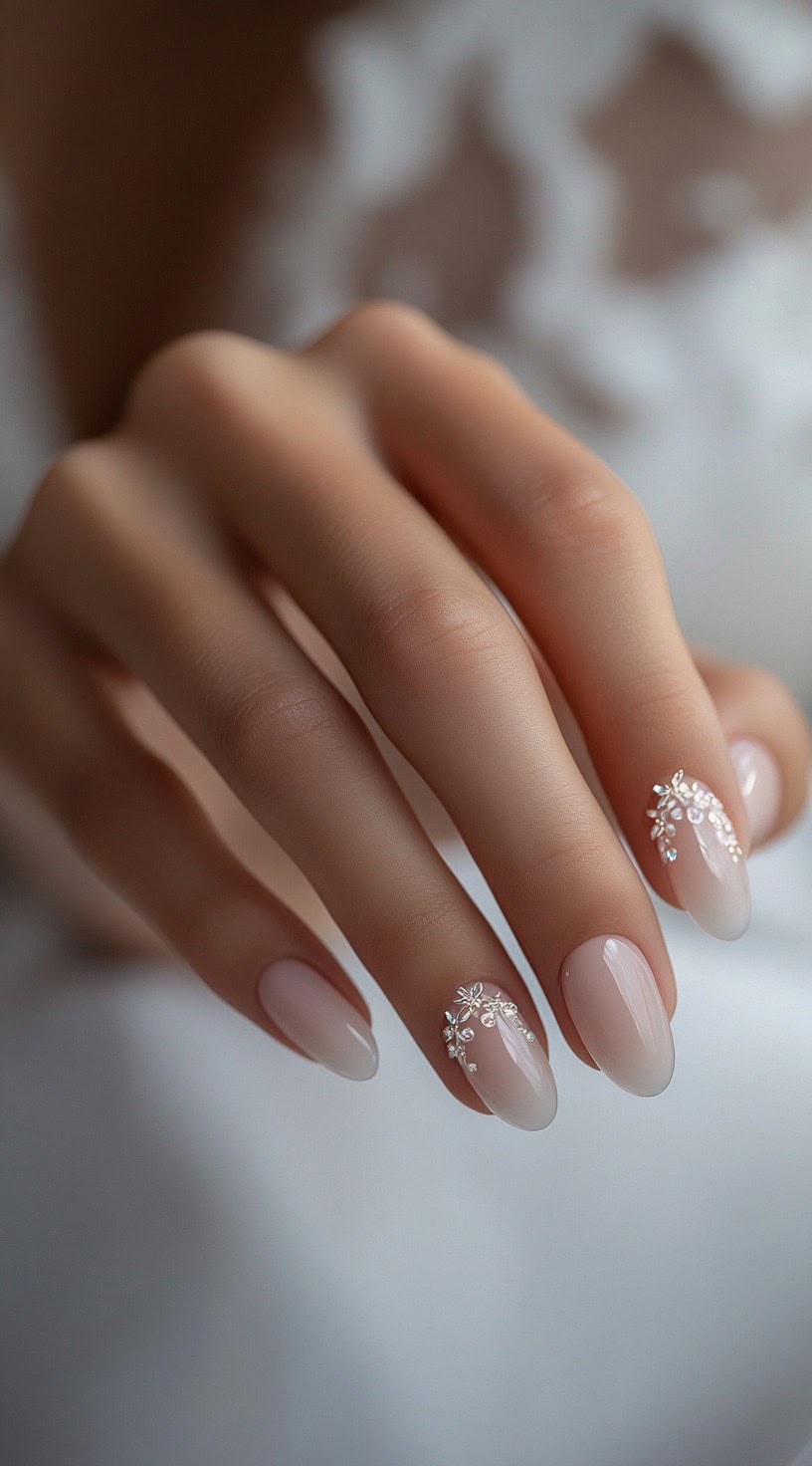 A hand with almond-shaped nails featuring a soft nude ombre base with delicate floral embellishments and tiny rhinestones on select nails.