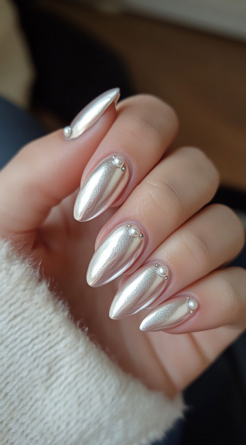 A close-up of almond-shaped nails with a soft silver chrome finish, each adorned with a delicate pearl accent near the cuticle.