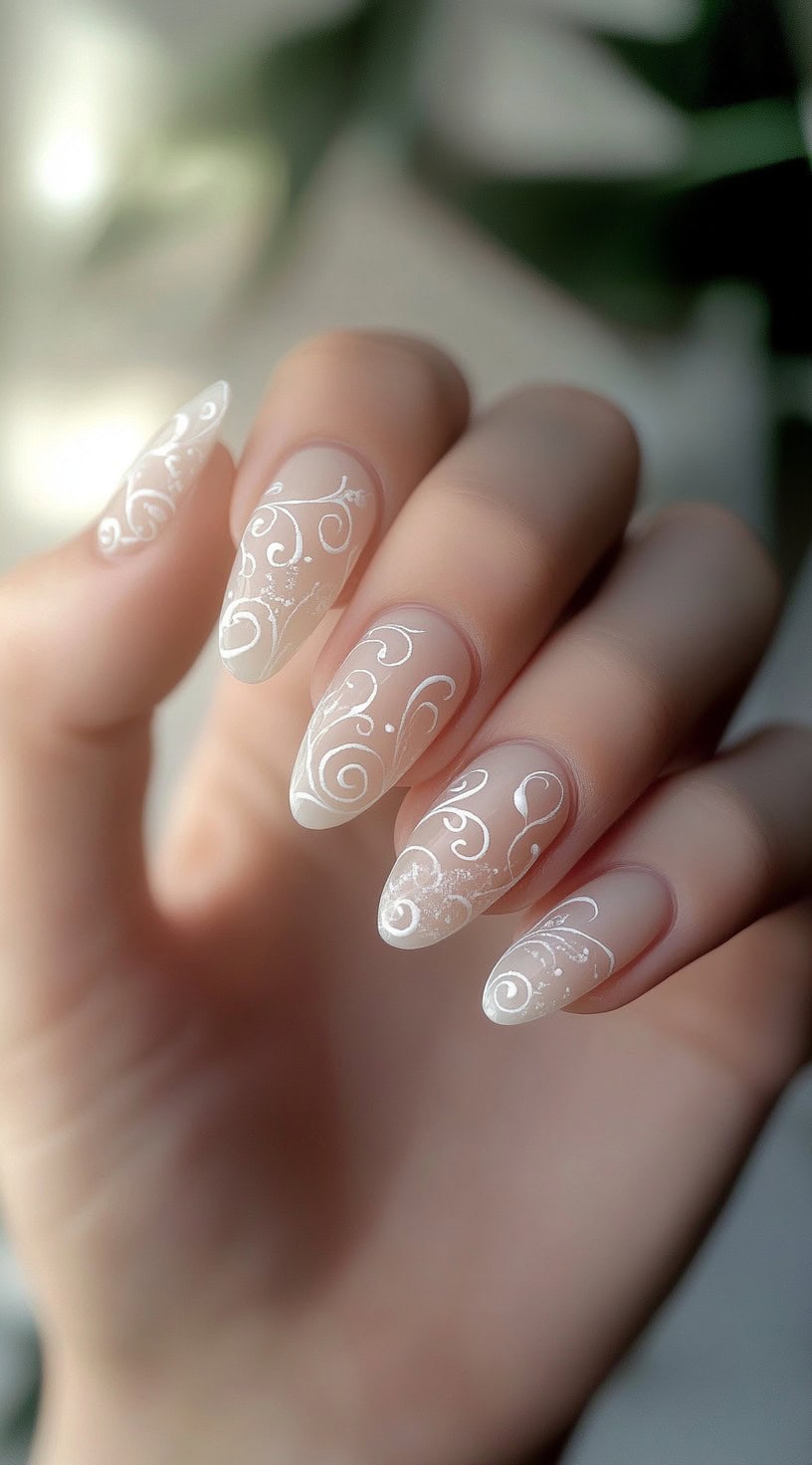 A hand with almond-shaped nails featuring a soft nude base and intricate white swirl designs, creating a delicate, lace-like effect.