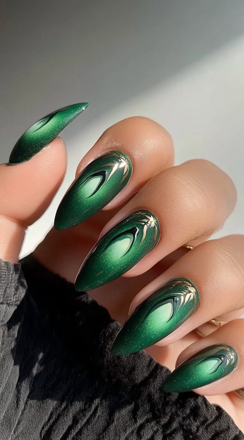 A hand with almond-shaped nails featuring a deep green cat-eye effect with gold metallic accents near the cuticle.