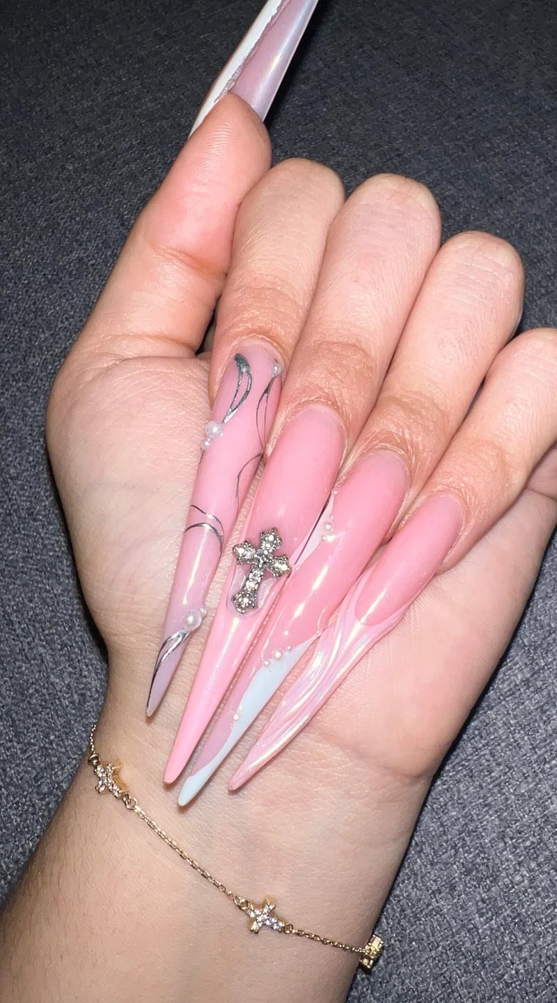 Extra-long pink stiletto acrylic nails with a glossy finish, black swirl accents, and a rhinestone-studded cross charm.