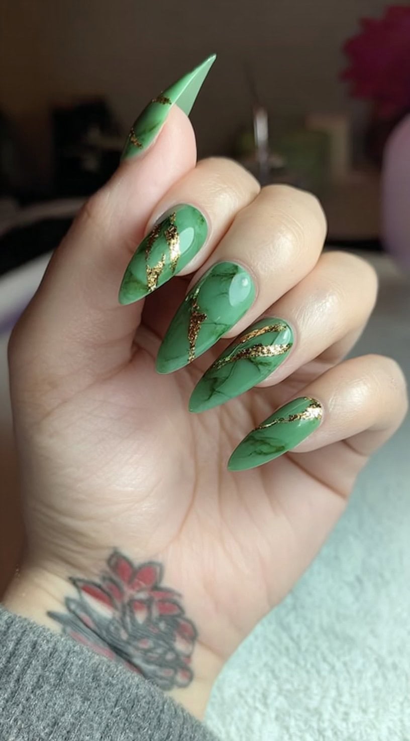 A hand with almond-shaped nails painted in rich green jade with gold foil veining that mimics cracked marble.