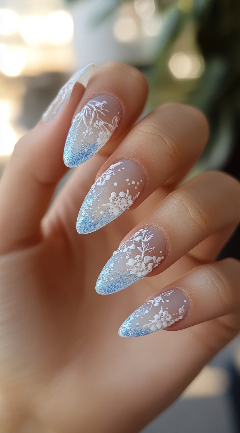 A close-up of stiletto-shaped nails with sheer frosted tips, delicate white floral designs, and shimmering blue glitter accents.