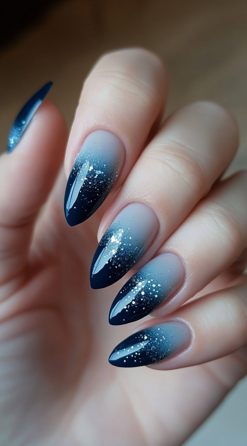 A hand with almond-shaped nails featuring a soft frosted ombre fading into dark blue glitter tips.