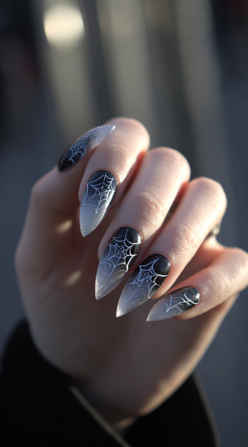A hand with stiletto nails in black-to-clear frosted ombre, each decorated with sharp white spider webs.