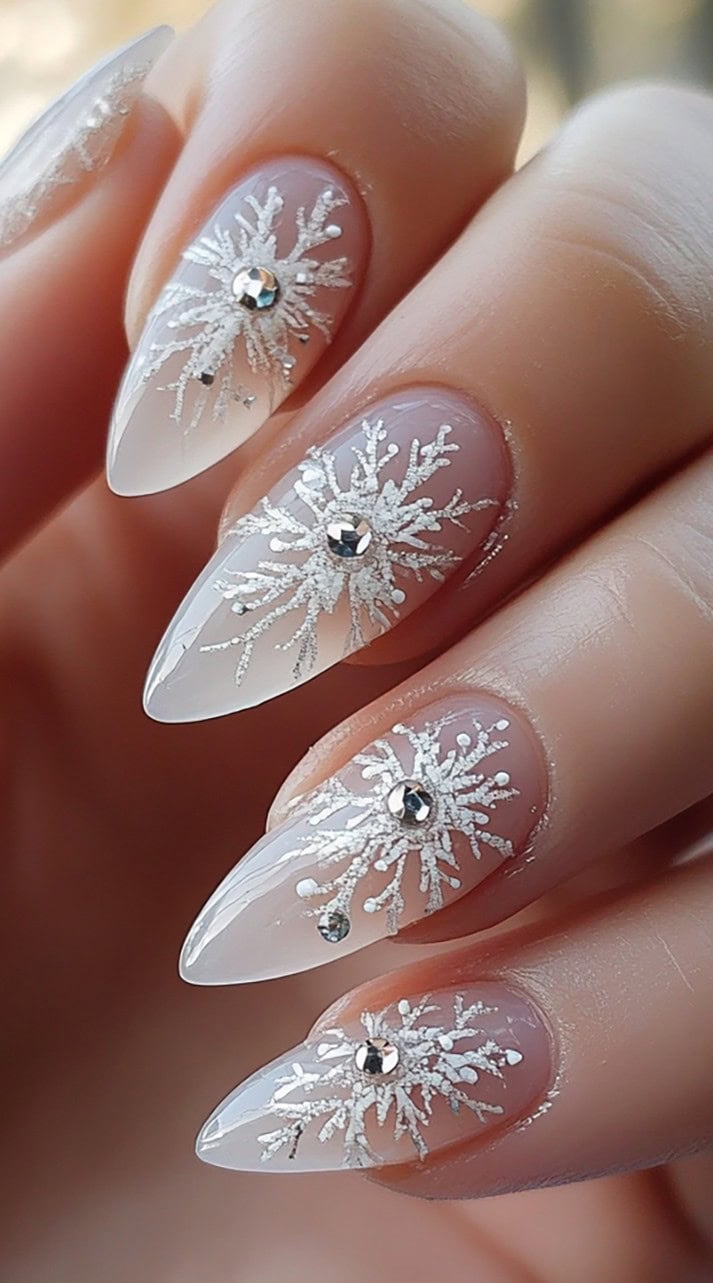 A close-up of almond-shaped nails with a sheer nude base, adorned with intricate silver glitter snowflakes and rhinestone embellishments.