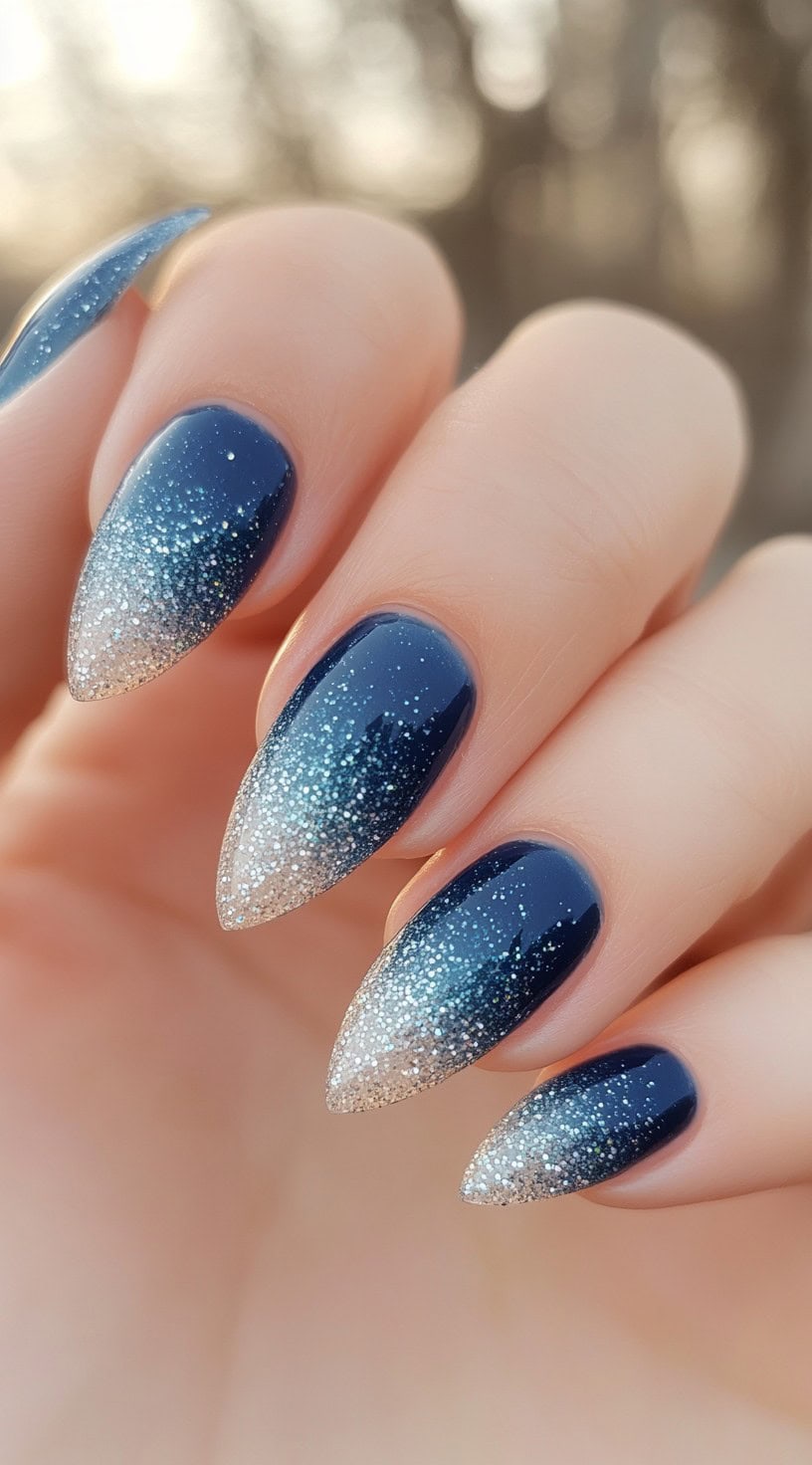 Pointed nails with navy blue to silver-white ombre glitter fading toward the tips.