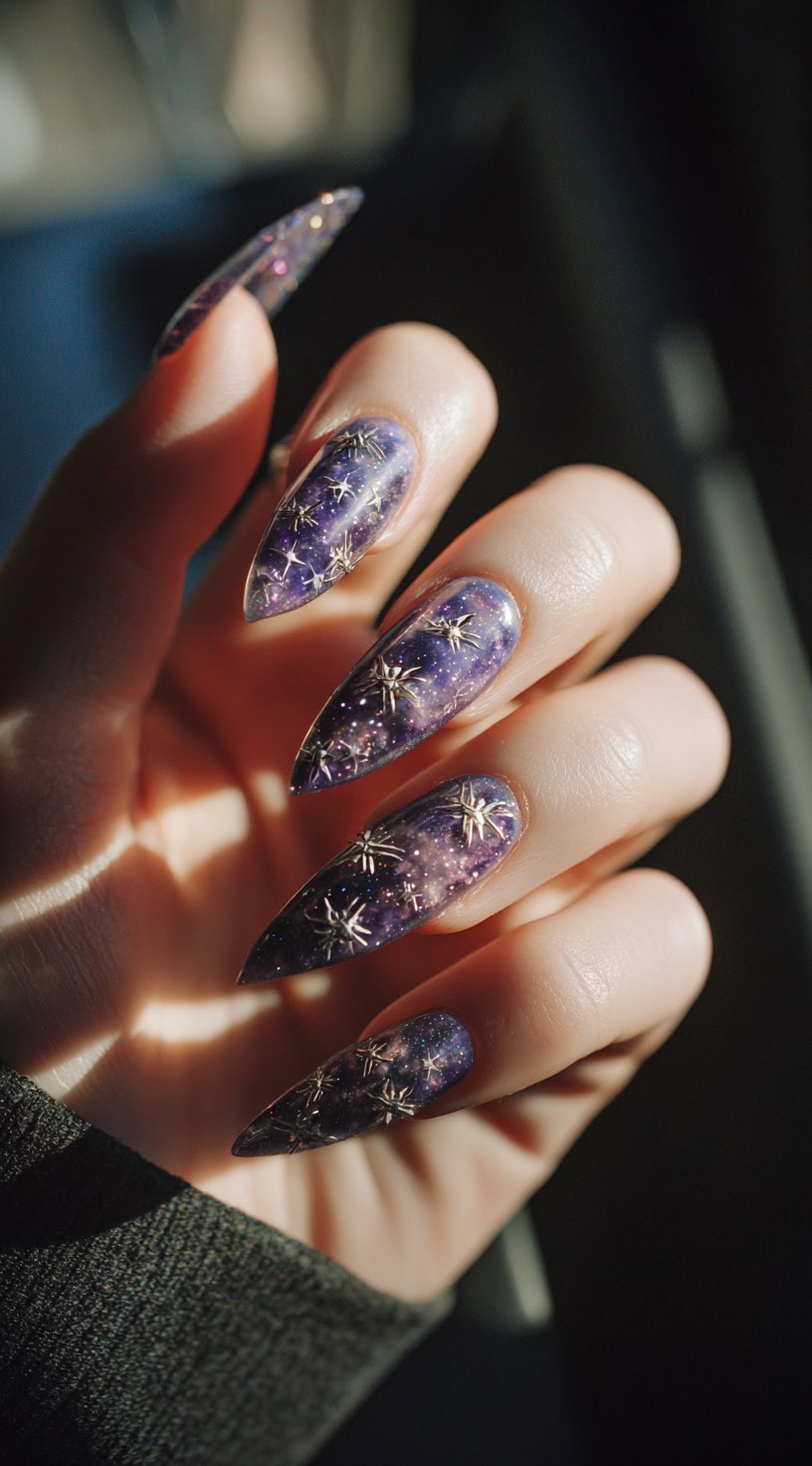 A hand with long stiletto nails featuring a purple galaxy background and raised silver spider charms.