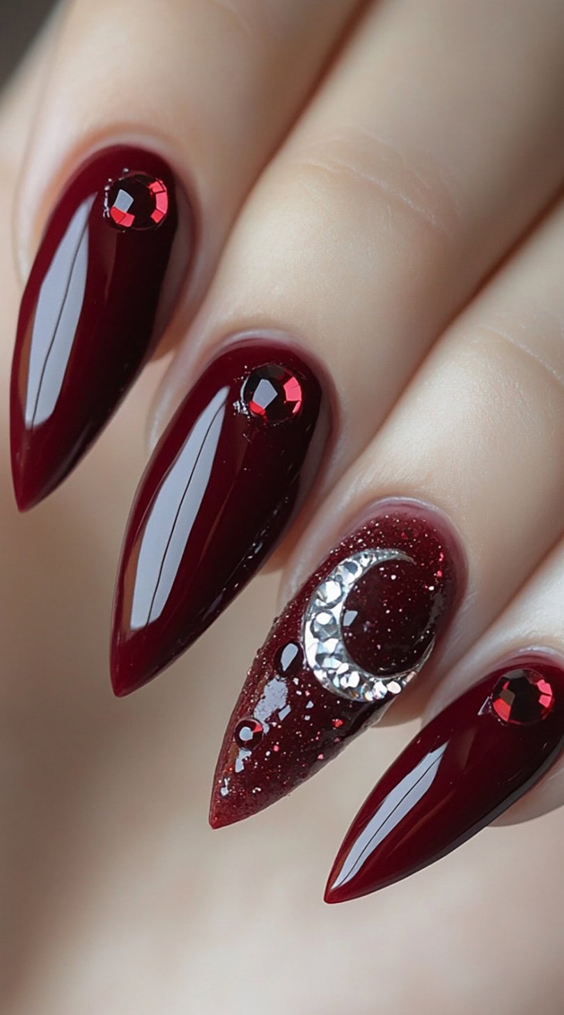 Garnet-toned almond nails featuring glossy red polish with red gems and a crescent moon made of rhinestones.