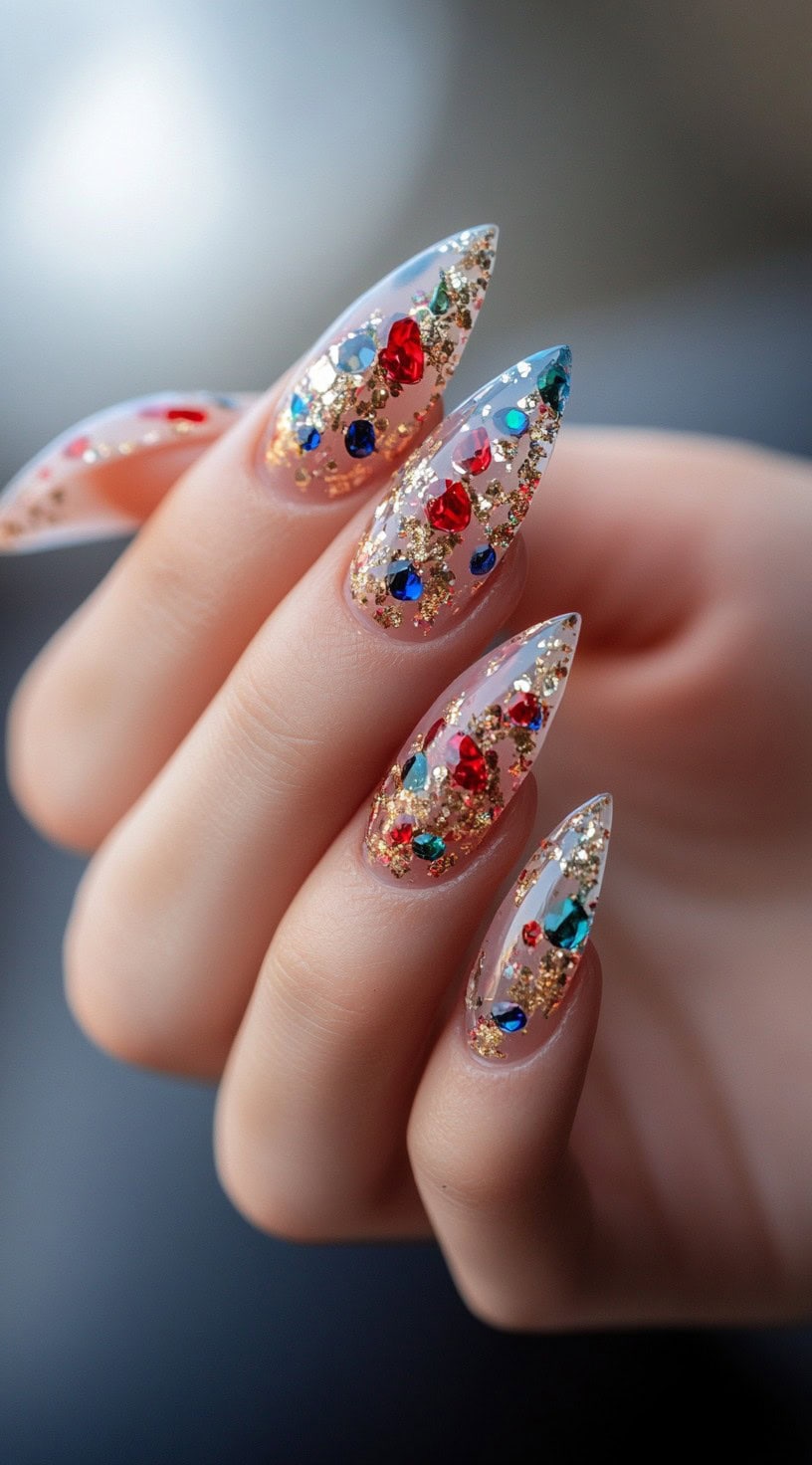 A hand showcasing long, stiletto nails with clear tips embedded with red and blue gemstones and gold glitter.