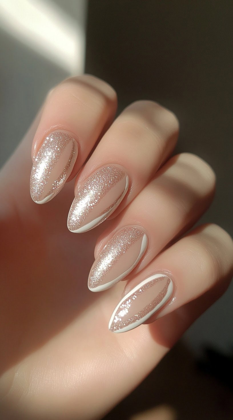 A hand with almond-shaped nails featuring a nude base with glittery gold tips and delicate white line detailing.