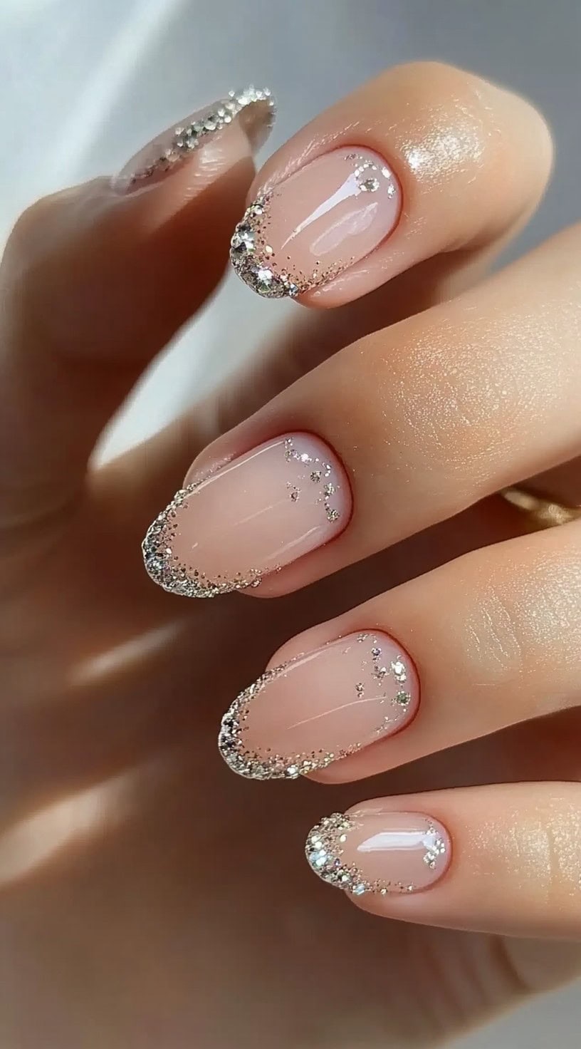 A close-up of a hand with short almond nails featuring a sheer pink base and glittery silver tips, creating a sparkling gradient effect.