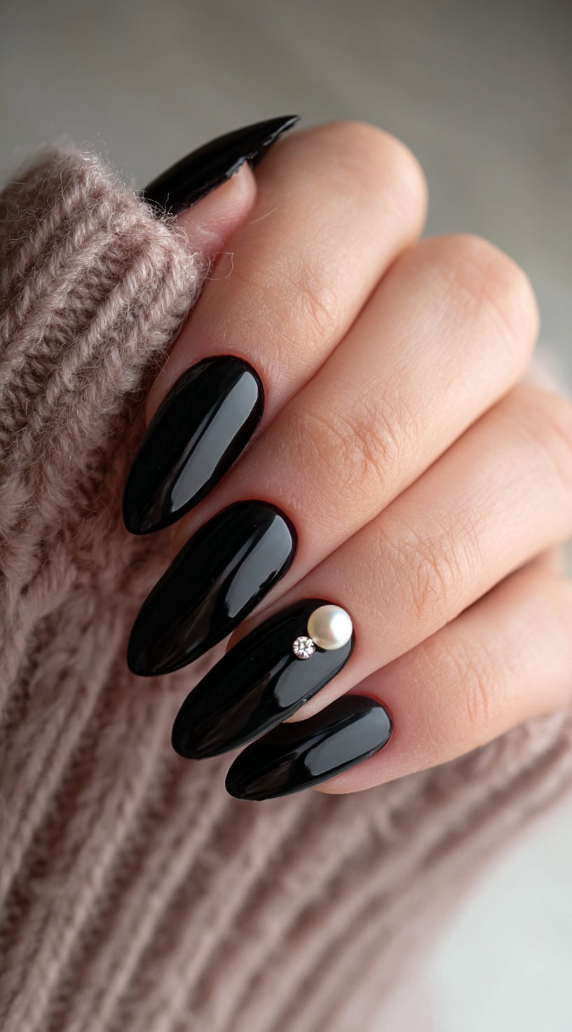 Glossy black almond nails, with one nail decorated using a small pearl and a rhinestone.