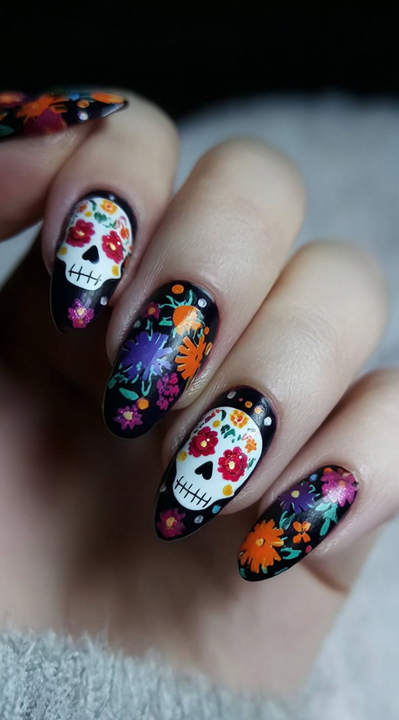 Hand with almond-shaped glossy black nails featuring white sugar skulls and bright floral designs.