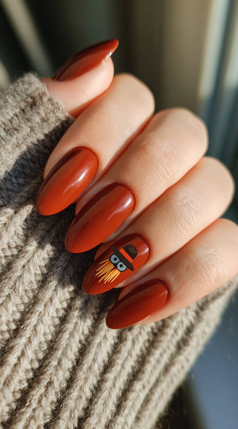 Glossy almond-shaped nails in burnt brown with a small cartoon scarecrow face wearing a hat on the ring finger.