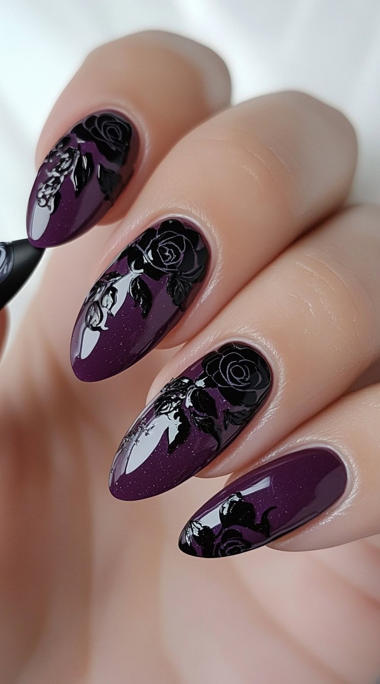 A hand with almond-shaped nails in deep purple polish, accented with glossy black rose designs and subtle shimmer.
