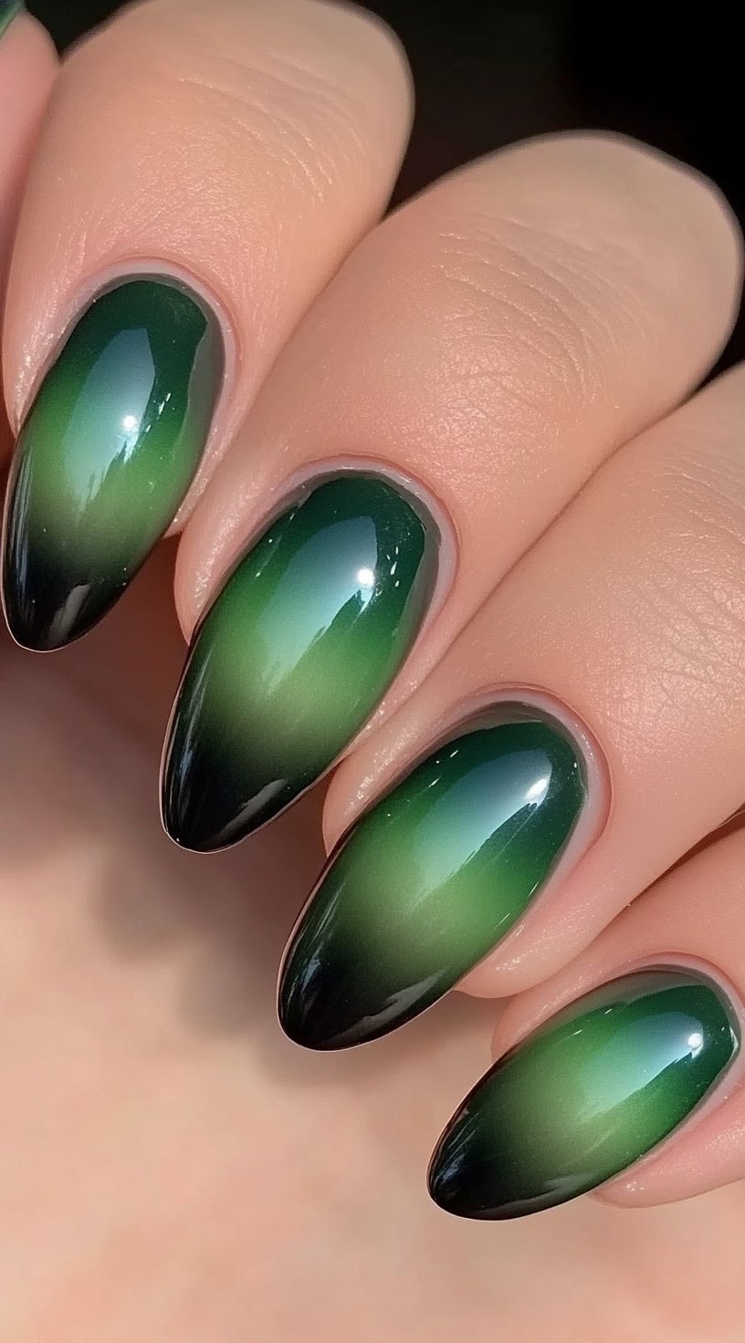 A close-up of a hand with almond-shaped nails featuring a seamless gradient from deep green to black with a glossy finish.