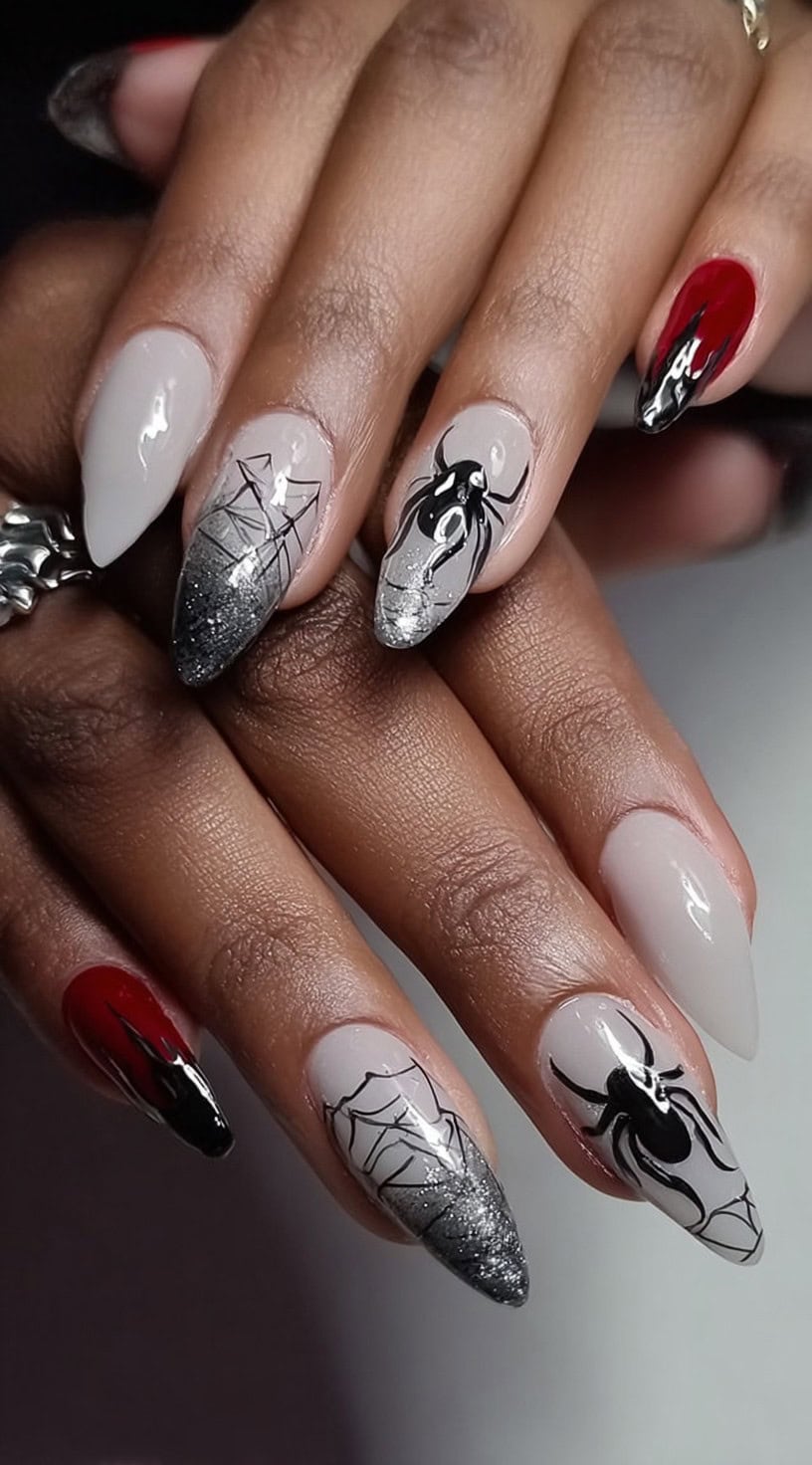 A hand with almond-shaped nails in taupe and red, featuring black web art, black 3D spiders, and silver glitter gradients.