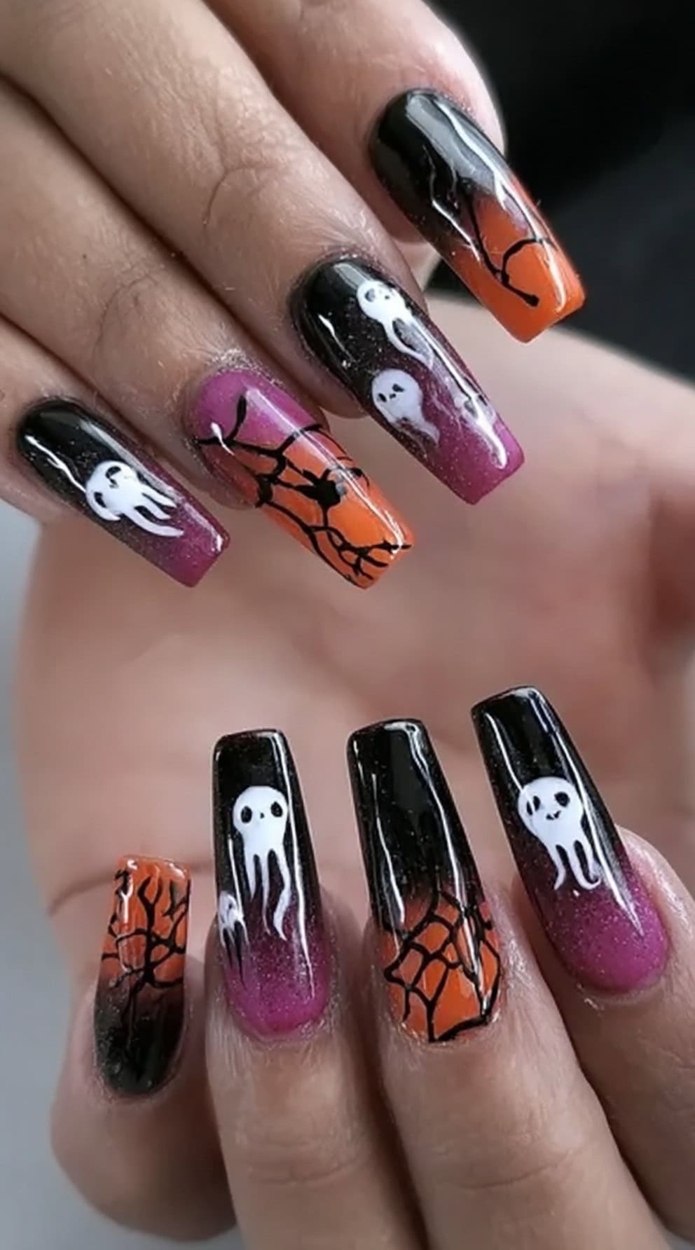 A hand with long square nails in black, orange, and pink ombré featuring cartoon ghosts and web patterns.