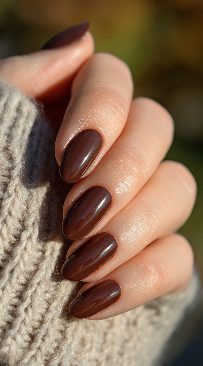 A close-up of oval-shaped nails painted in a glossy chocolate brown polish, paired with a beige knit sweater.