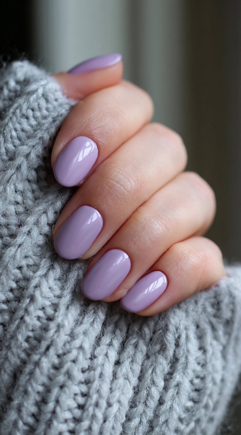 Oval-shaped nails painted in a glossy pastel lilac color, shown against a grey knit sweater.