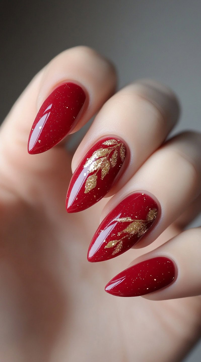 A set of glossy red almond-shaped nails with delicate gold leaf detailing placed in an organic, hand-painted pattern.